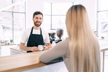 A young barista in a black apron smiles as he brings a cup of co