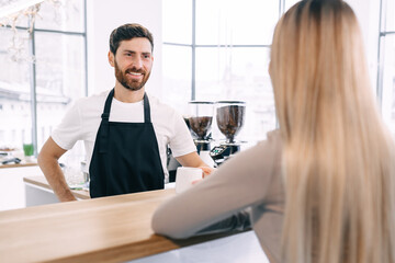 A young barista in a black apron smiles as he brings a cup of co