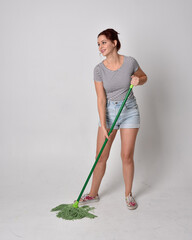 full length portrait of girl wearing shirt and denim shorts, holding a broom. isolated on grey studio background.