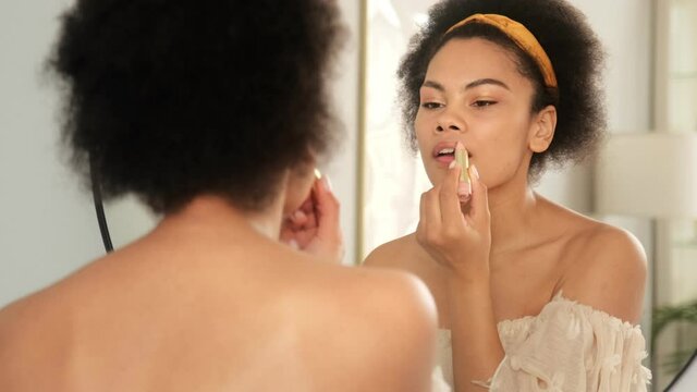 Black african american woman doing make up, holding lipstick and paints lips. She looking at mirror reflection and smiles. Get ready in morning.