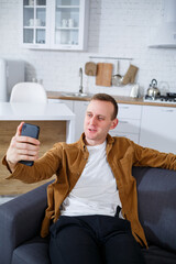 A young successful man is sitting at home on the couch with a phone and talking on a video link. Remote work during quarantine.