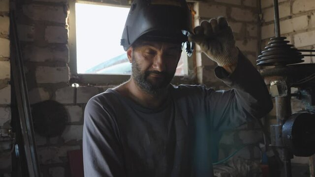 Portrait of adult welder opens protective mask after finishing his job and looking into camera in workshop. Professional mechanic with beard working in his garage. Close up Slow motion