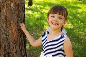A little beautiful girl in a dress stands near the trunk of a tree in the pine forest