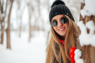 Young woman in sunglasses and hat hiding behind the tree in a snowy park
