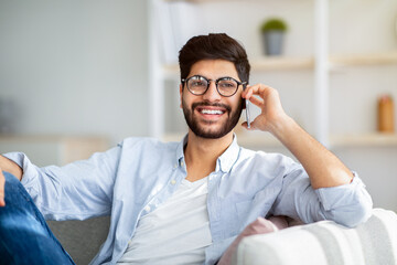 Phone conversation concept. Cheerful arab man talking on smartphone resting on sofa at home interior