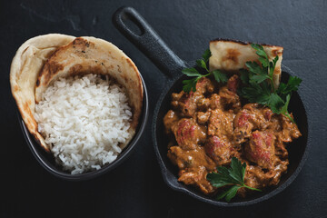 Cast-iron pan with indian chicken makhani, bowl of white rice and flatbread, high angle view over black stone background, studio shot