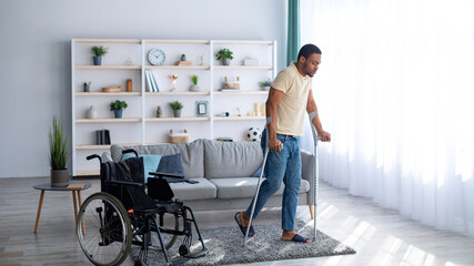 Rehabilitation concept. Young black guy walking with crutches after using wheelchair at home,...