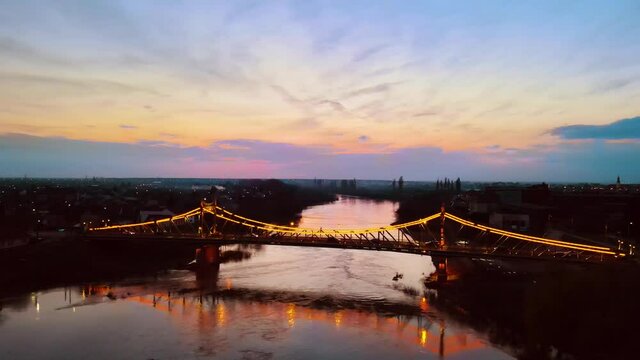 Aerial footage of a spanning bridge over a river, shot from a drone, while panning the drone from right to left keeping the bridge in focus. Drone video of an iluminated spanning bridge at sunset
