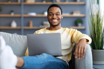Positive black man chilling on sofa, using laptop
