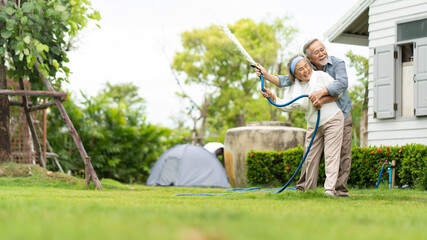 Asian senior couple smiling and watering green cultivated plants with a garden hose at home. Happy...