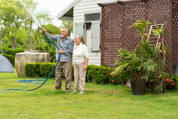 Asian senior couple smiling and watering green cultivated plants with a garden hose at home. Happy...