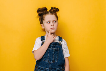 Thoughtful little preschool girl child touch chin with finger thinking or considering, pensive lovely small kid making decision imagining idea, isolated over yellow studio background with copy space