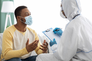 Doctor in PPE suit taking anamnesis for sick male patient - obrazy, fototapety, plakaty