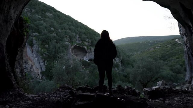 Slowmotion of a girl standing inside a cave observing the beautiful landscape background. The camera moving forward towards her back.