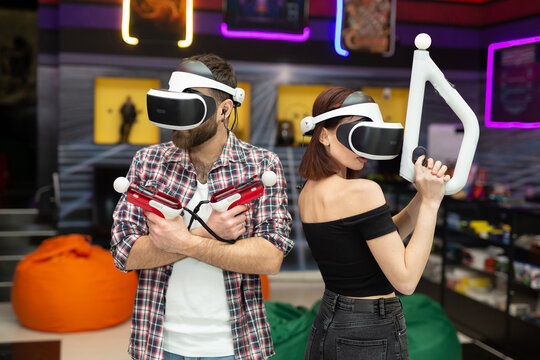 Friends, A Man And A Woman Use A Virtual Reality Headset With Glasses And Hand Motion Controllers And Weapons In The Play Area.