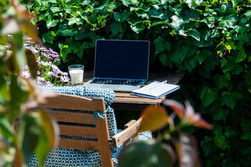 Remote workplace, laptop, notebook with a pen, a glass of milk are on a wooden table, next to a chair with a blue pillow, all this is in the green garden with blurred leaves, not the foreground