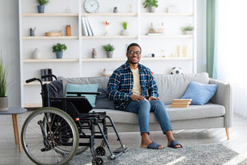Happy disabled black guy sitting on sofa with books, empty wheelchair standing nearby, full length...