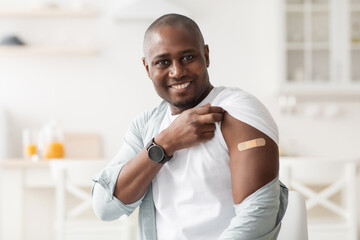 Vaccination concept. Happy african american man holding up shirt sleeve showing plaster after...