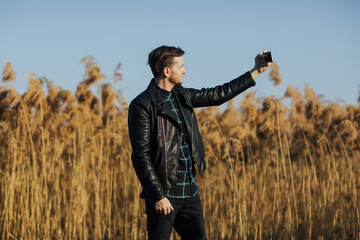 Stylish man in a black leather jacket stands against a background of fluffy reeds and blue sky on a sunny day and taking selfie.