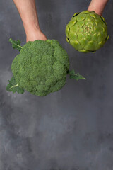 Caucasian male hands holding broccoli and green artichoke isolated on gray background.