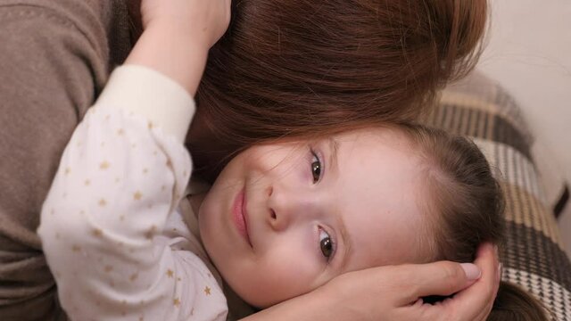 Little happy girl hugs her beloved mother and kisses, the child with the mother lie on the sofa in the children's room and laugh, parental affection and support for the kid, the concept of family