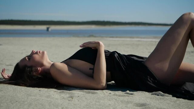 View from the side of an attractive girl with a beautiful figure in a black dress posing for the camera lying on the sand by the river in clear sunny weather. High quality FullHD footage