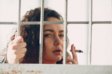 Young brunette curly woman in orange suit behind jail bars smokes marijuana joint. Female in...