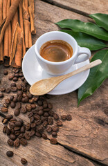 Espresso in a white mug, coffee beans in a wooden bowl and cinnamon sticks placed on a wooden table.