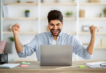 Overjoyed arab entrepreneur celebrating business success with raised arms, looking at laptop screen...