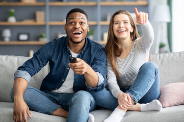 Cheerful multiracial couple having fun at home, watching comedy movie on tv - obrazy, fototapety, plakaty
