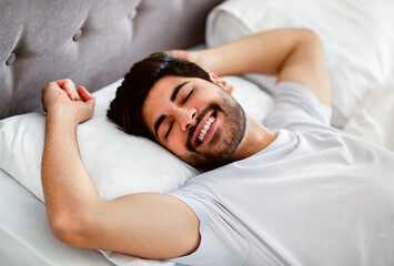 Fototapeta na wymiar Happy wellslept arab man awakening and stretching hands lying in modern comfy bed at home
