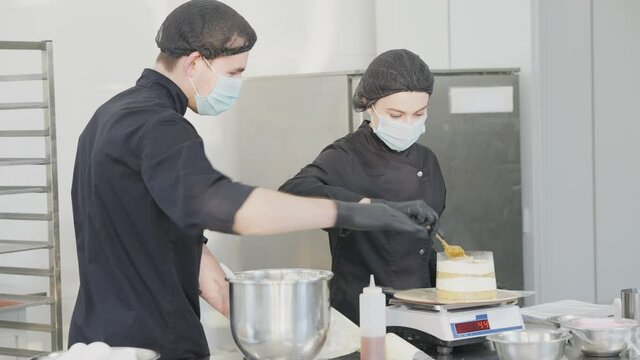 Side view of professional man teaching woman cooking dessert in commercial kitchen. Confident Caucasian confectioner in Covid face mask controls trainee in candy store on coronavirus pandemic.