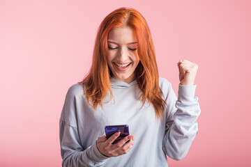 Fototapeta premium Redhead girl looks at the phone and shows the gesture of the winner in the studio on a pink background