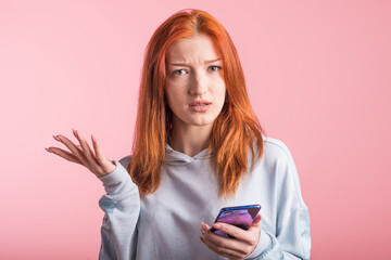 Fototapeta premium Confused redhead girl with phone in studio on pink background