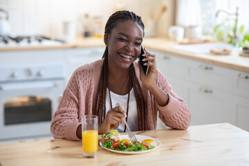 Cheerful African American Female Eating Breakfast And Talking On Cellphone In Kitchen