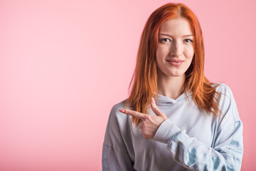 Redhead girl showing index finger to the side for copyspace in studio on pink background