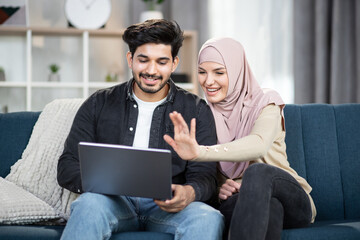 Smiling happy muslim couple witl laptop computer at home, waving hands while having video call with...