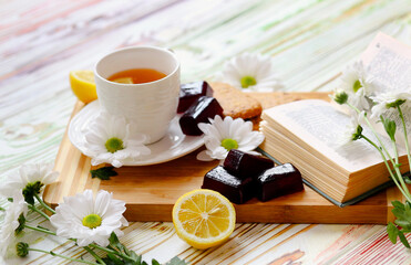 Banner with a breakfast: tea, sweets, lemon, marmalade on a beautiful light wooden background and decorate are book and flowers. Sweets. Sunlight from the window. Morning concept