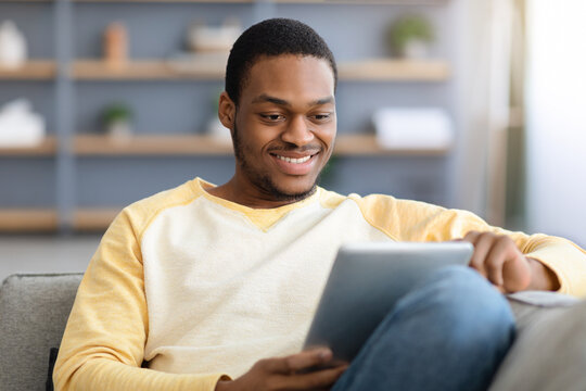Smiling afican american guy sitting on couch, using digital tablet