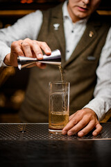 bartender holds steel jigger and carefully pours liquor into glass on the bar