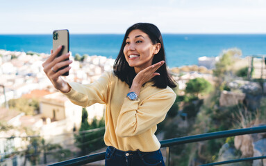 Asian female vlogger grinning while clicking selfie images via cellular front camera using roaming...