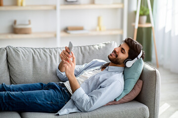 Free time. Relaxed arab guy listening to music in wireless headphones, lying on comfy couch