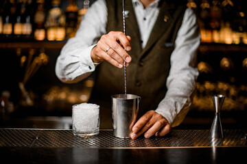 Fototapeta na wymiar view on bartender holding long bar spoon and stirring cocktail in steel mixing cup