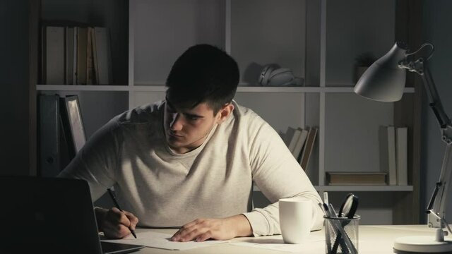 Night learning online. Internet meeting. Distance education. Smart male student working late at virtual class using laptop listening watching video lesson writing taking notes at dark workplace.