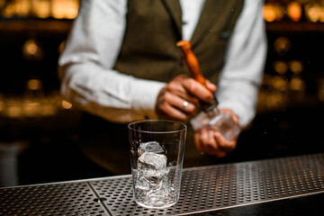 glass with ice cubes on bar. Bartender is cracking ice in the background