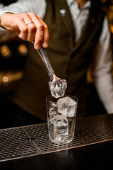bartender holds piece of ice with tongs over glass on bar counter
