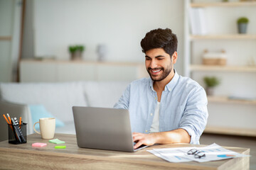 Positive freelancer working on laptop at home office, typing on keyboard and looking at screen