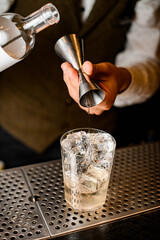 view of crystal glass with ice cubes in which bartender neatly pours drink from jigger