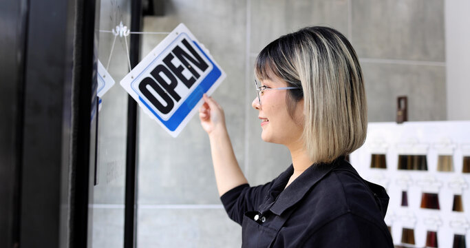 Young Asian Woman Business Owner Standing And Turning Open Sign On Glass Door Of Her Salon Shop