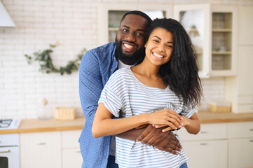 Obraz premium Happy married multiracial couple posing for a photo on kitchen, the happiest hearts make the happiest homes, african husband hugs latin wife with thick curly hair from behind, harmonic relationships